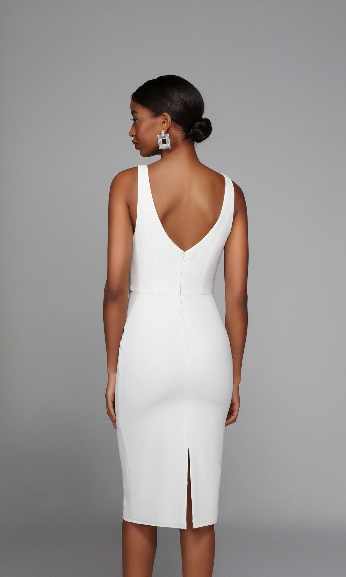 Woman wearing the knee-length semi-formal dress Alyce 70010, featuring a sleeveless white design, deep V-back, and back slit, stands against a plain gray background facing away from the camera.